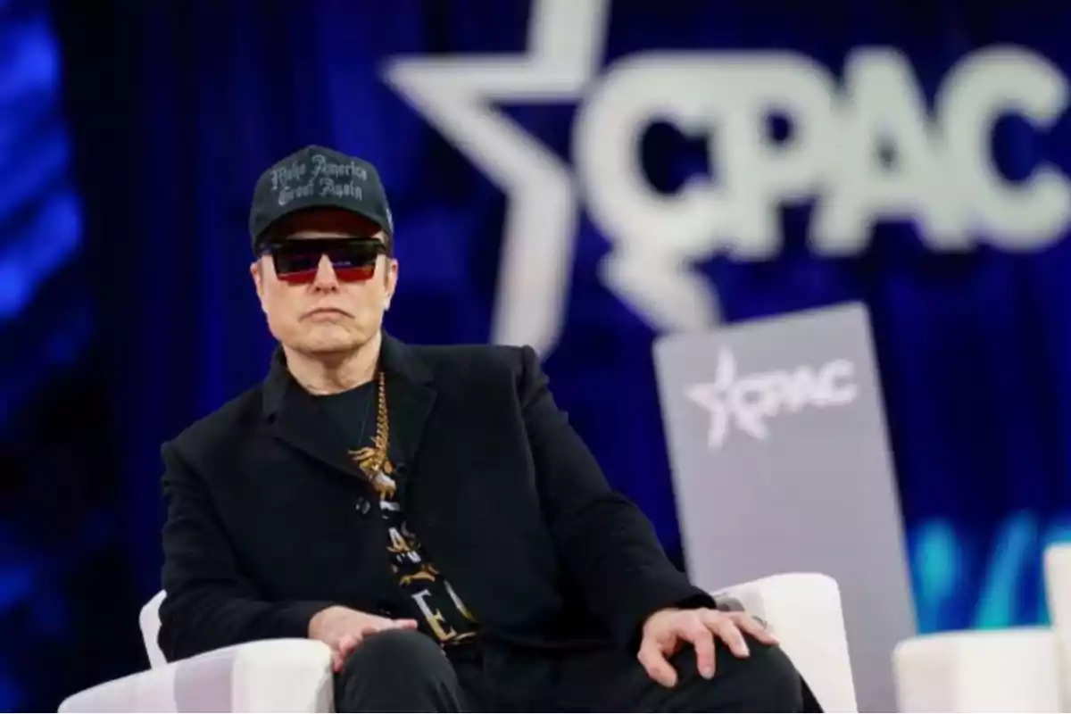 Un hombre con gafas de sol y gorra negra está sentado en un escenario con el logo de CPAC de fondo.