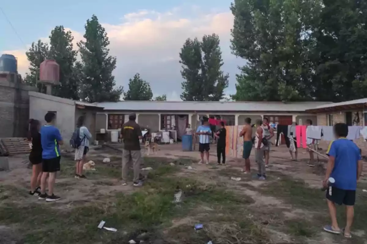 Un grupo de personas se encuentra reunido en un patio al aire libre frente a un edificio de una planta con árboles al fondo.