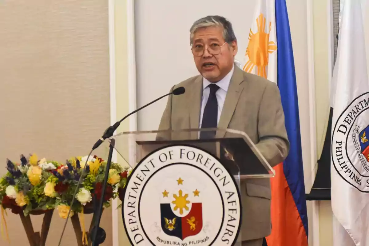 Un hombre de traje habla en un podio con el emblema del Departamento de Asuntos Exteriores de Filipinas y banderas de fondo.