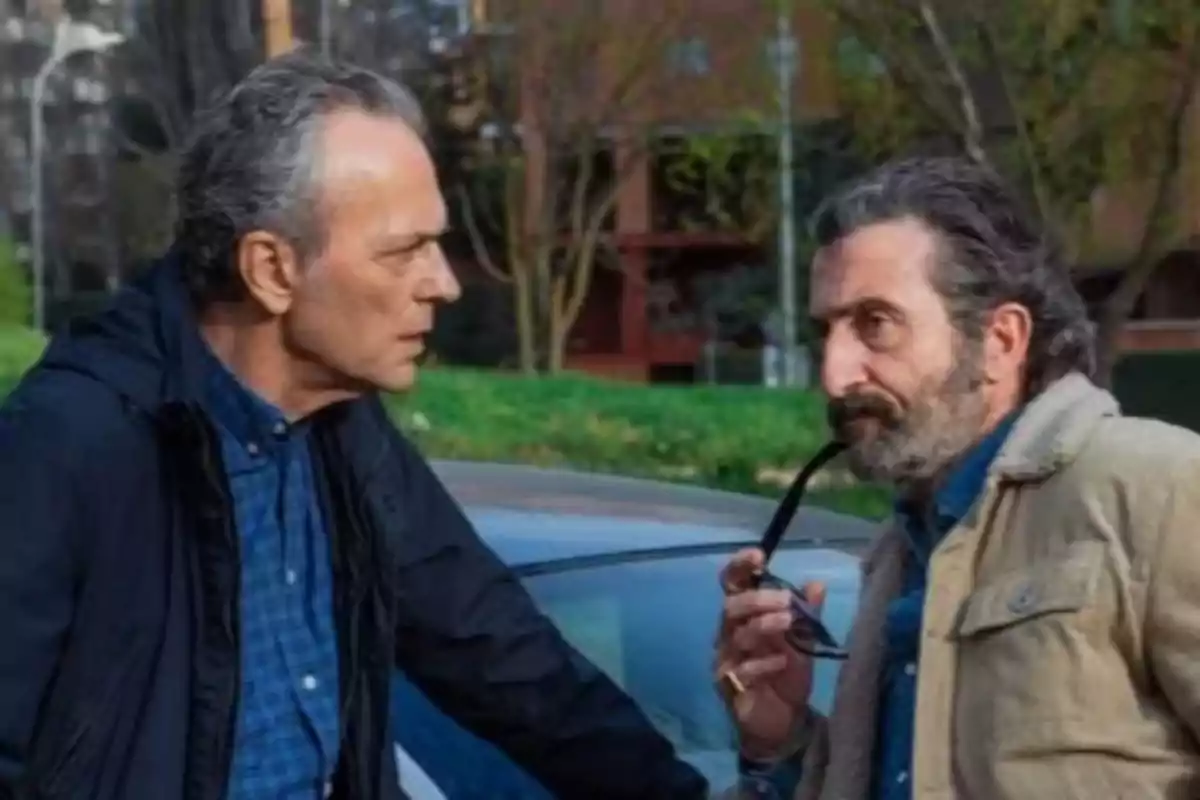 Dos hombres conversan al aire libre junto a un coche, uno de ellos sostiene una pipa en la serie 