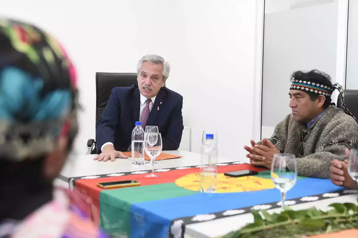 Un hombre de traje y corbata está sentado en una mesa junto a otras personas, una de las cuales lleva una vestimenta tradicional y una cinta en la cabeza, con una bandera colorida sobre la mesa.