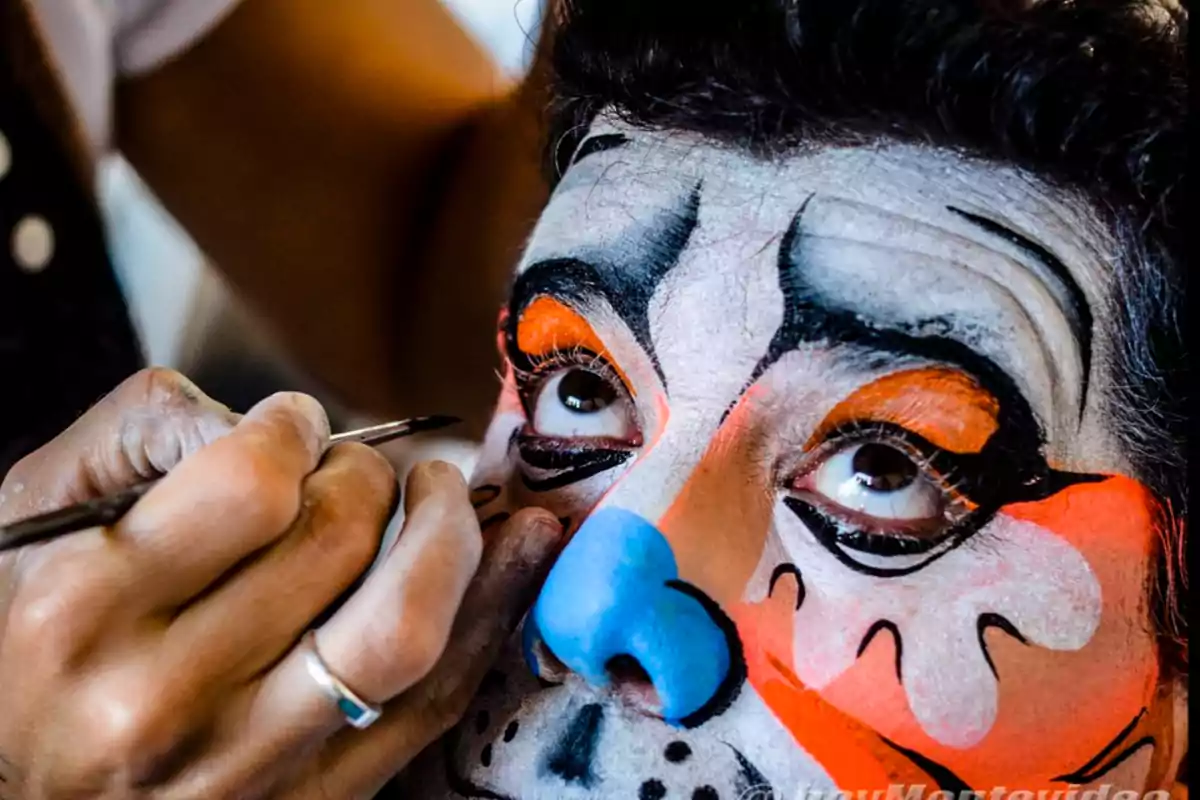 Persona con maquillaje colorido en el rostro mientras un artista aplica detalles con un pincel.