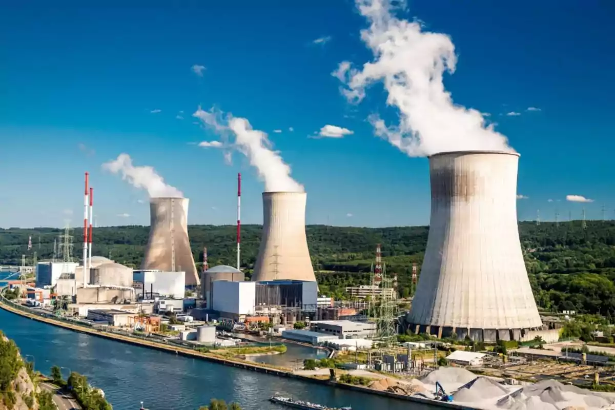 Planta de energía nuclear con torres de enfriamiento emitiendo vapor en un entorno natural con cielo azul.