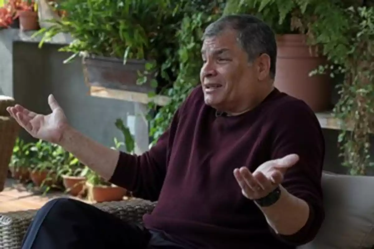 Hombre con suéter oscuro sentado en un entorno con plantas, levantando las manos en un gesto de incertidumbre.