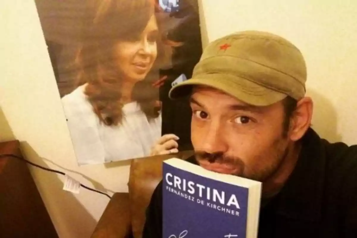 Un hombre con gorra sostiene un libro azul que tiene el nombre "Cristina Fernández de Kirchner" en la portada y detrás de él hay un póster de una mujer.