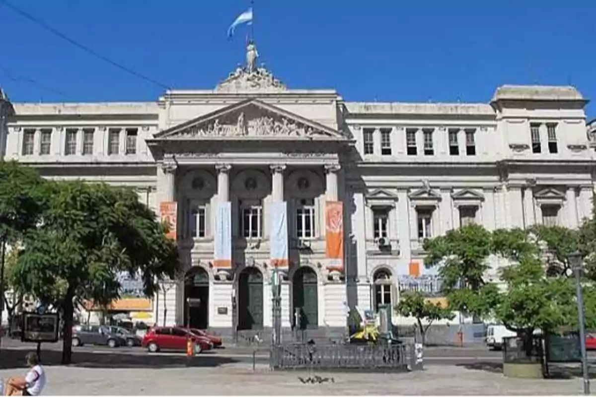 Edificio histórico con columnas y una bandera en la parte superior, rodeado de árboles y coches estacionados.