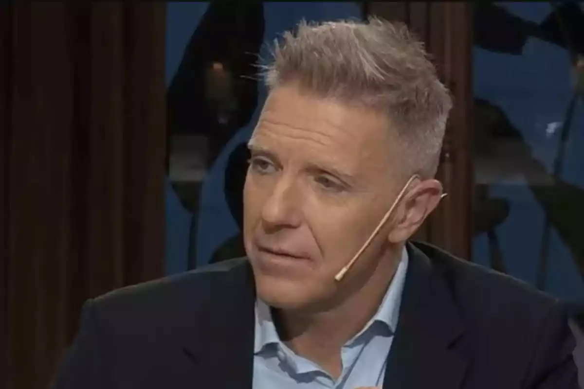 Hombre con cabello corto y canoso, usando un micrófono de diadema, vestido con camisa azul y saco oscuro, en un entorno de interior.