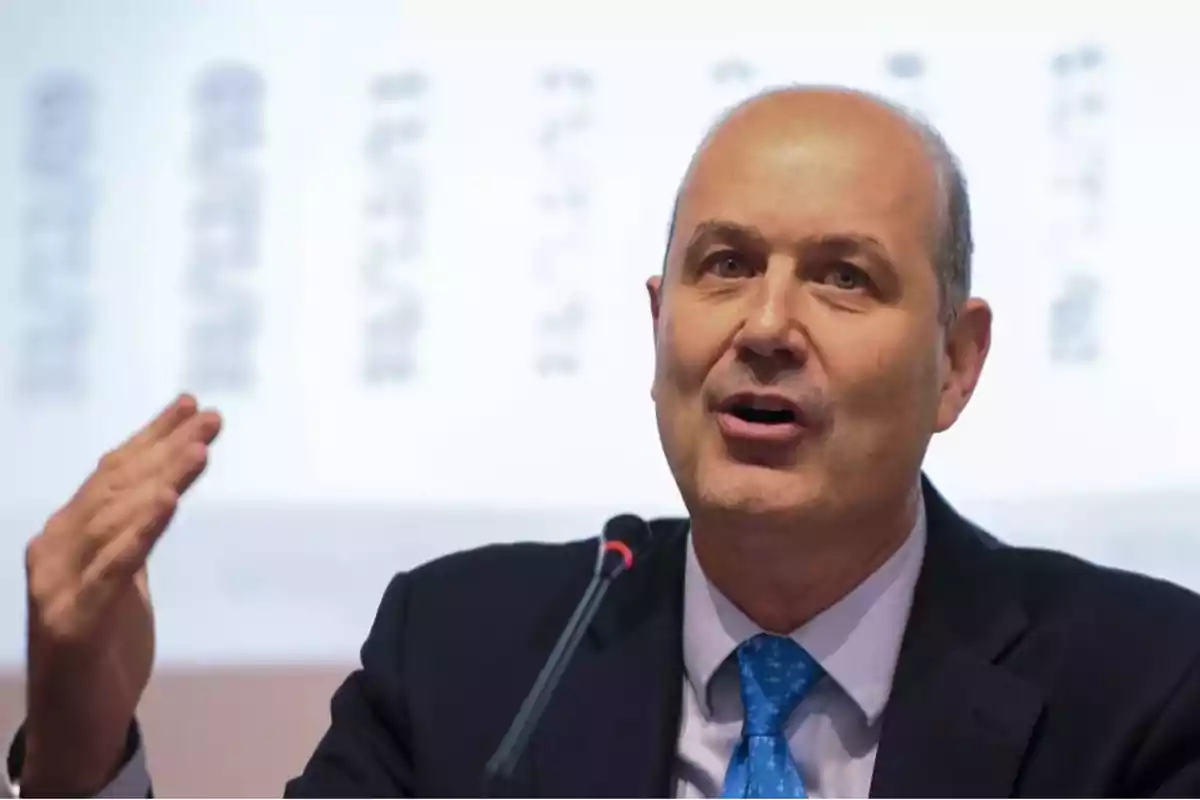 Un hombre hablando en una conferencia con un micrófono frente a él y una pantalla de fondo.