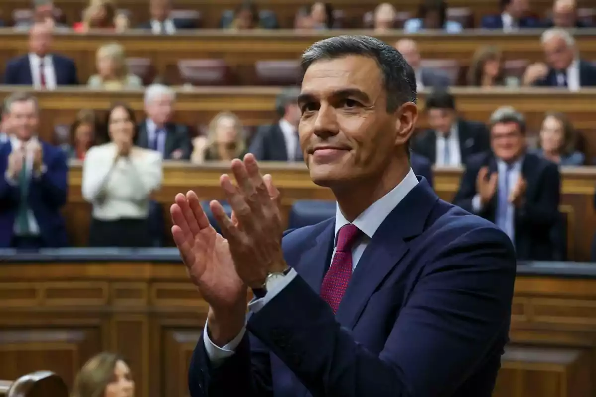 Un hombre aplaudiendo en un entorno formal con personas al fondo también aplaudiendo.