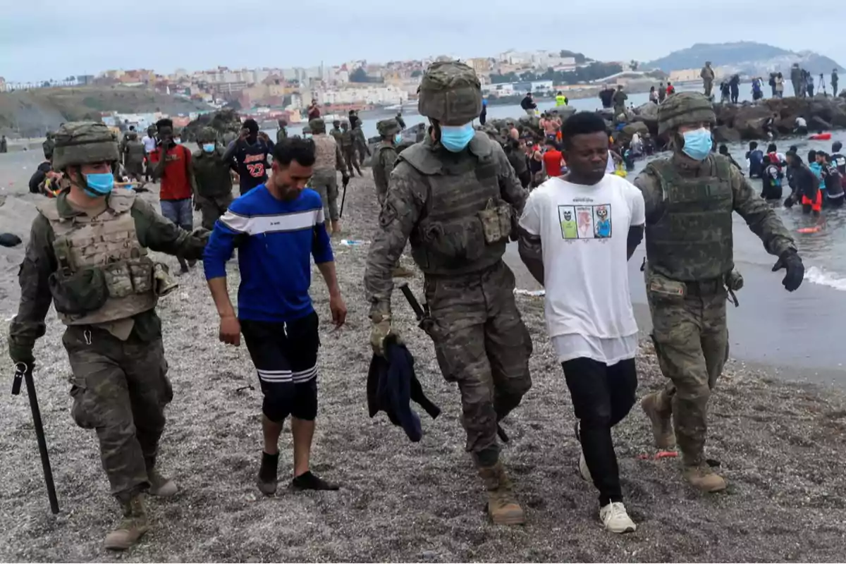 Soldados escoltan a dos hombres por una playa mientras otras personas están en el agua y en la orilla con una ciudad al fondo.