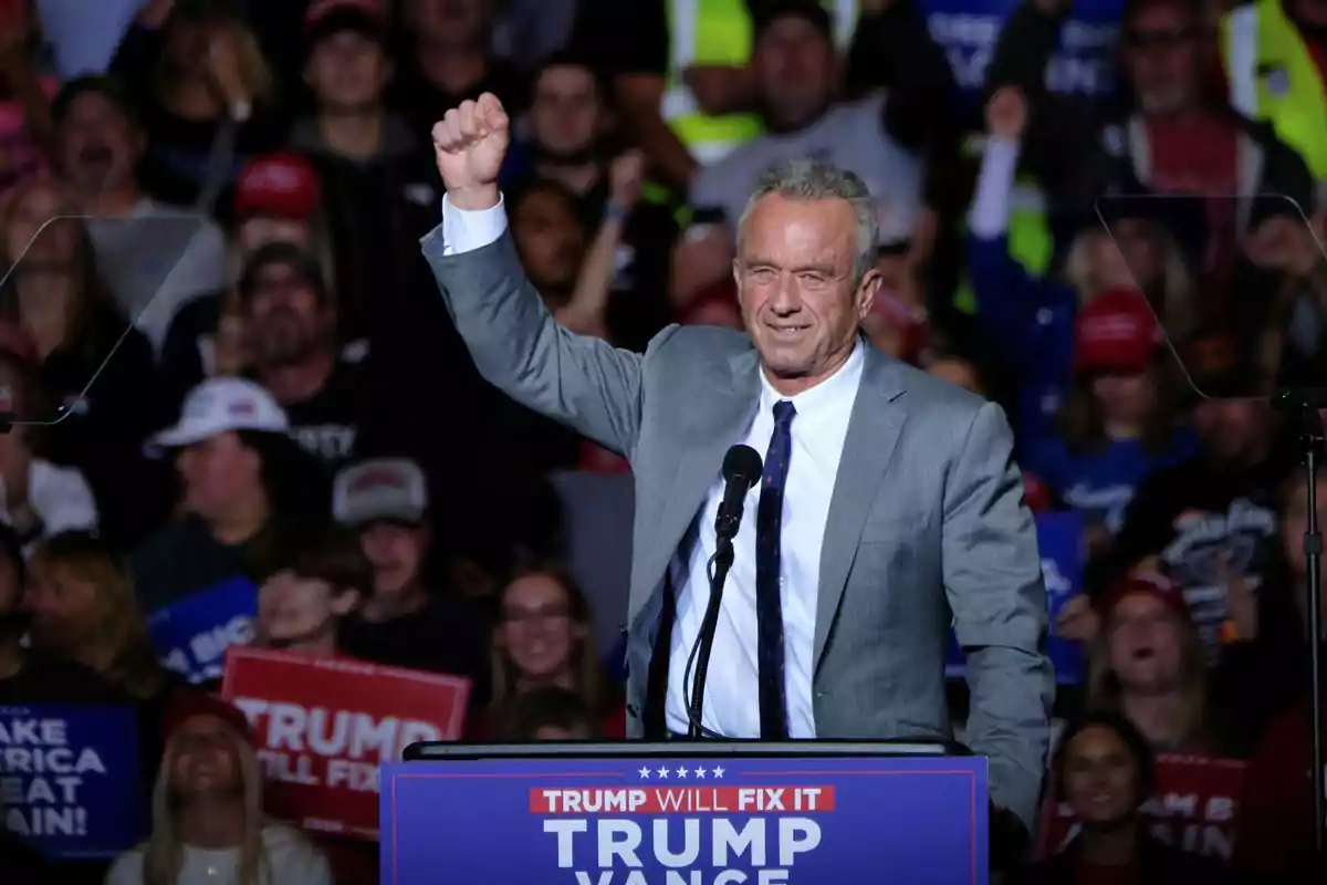 A man in a suit raises his fist while speaking at a political rally, surrounded by a crowd holding signs.