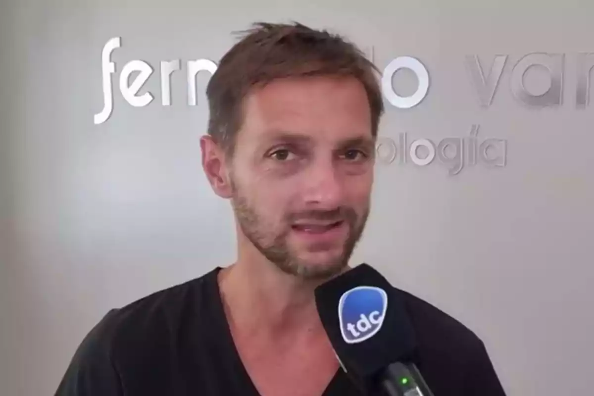 Un hombre con barba y cabello corto sostiene un micrófono con el logo 