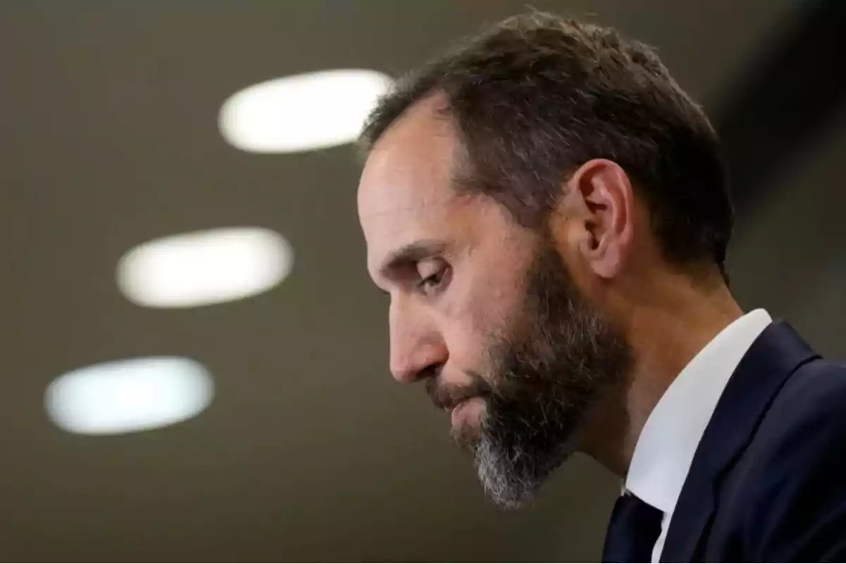 Un hombre de perfil con barba y traje oscuro en un entorno iluminado.