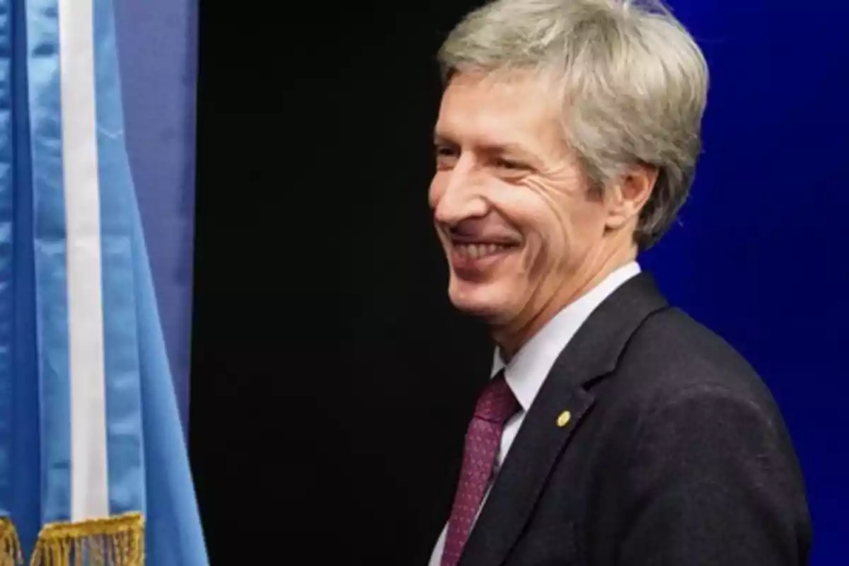 Hombre sonriendo de perfil junto a una bandera azul y blanca.