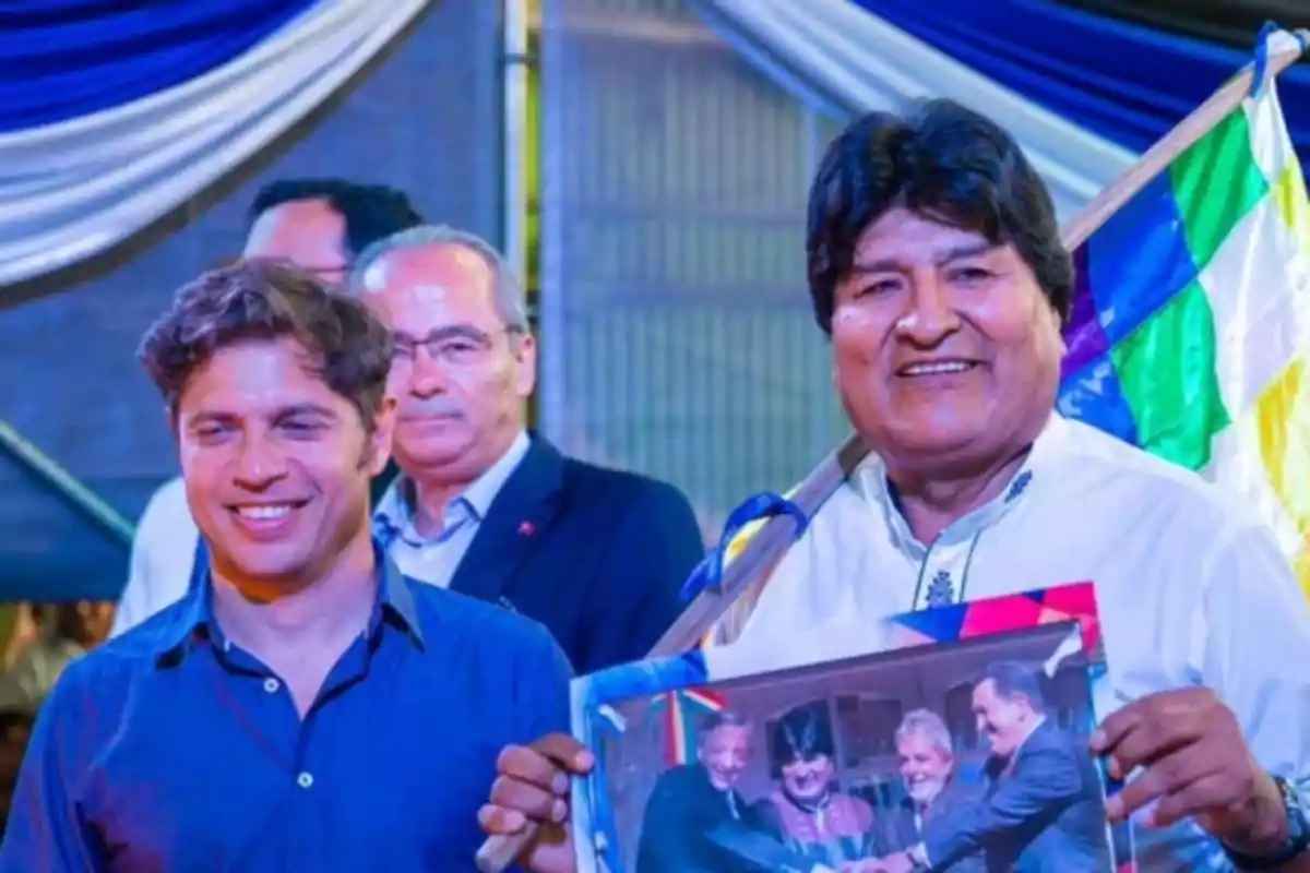 Un grupo de personas sonríe mientras una de ellas sostiene una bandera colorida y una fotografía.