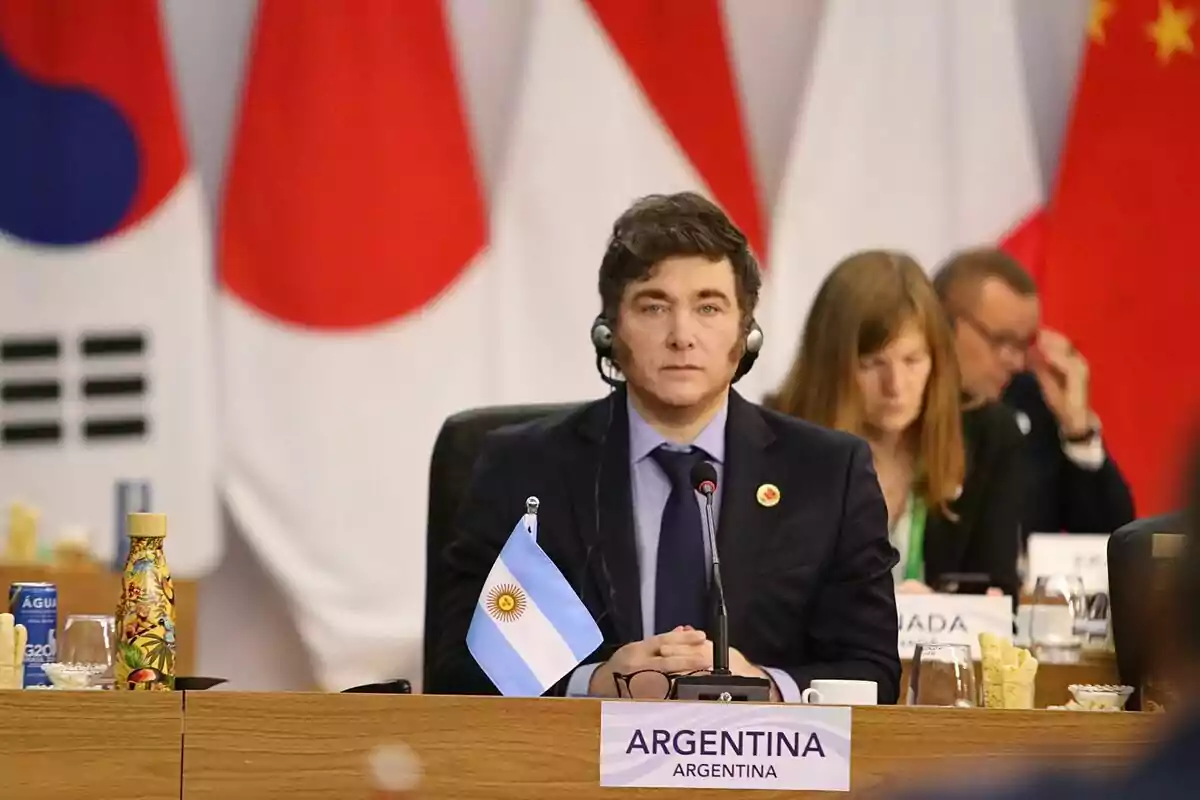 Un hombre con auriculares está sentado en una mesa de conferencia con una bandera de Argentina frente a él y banderas de otros países en el fondo.