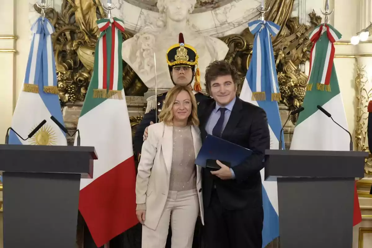 Dos personas posan juntas frente a banderas de Argentina e Italia en un entorno formal con un guardia detrás.