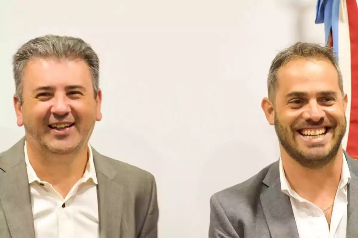 Dos hombres sonrientes vestidos con trajes claros posan frente a un fondo blanco.