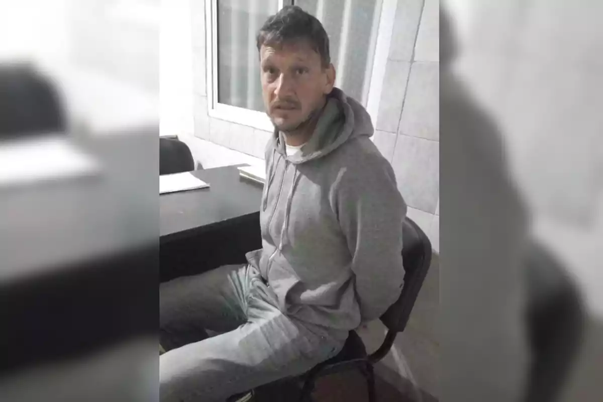 Un hombre con sudadera gris y jeans está sentado en una silla con las manos detrás de la espalda en una habitación con paredes de azulejos.