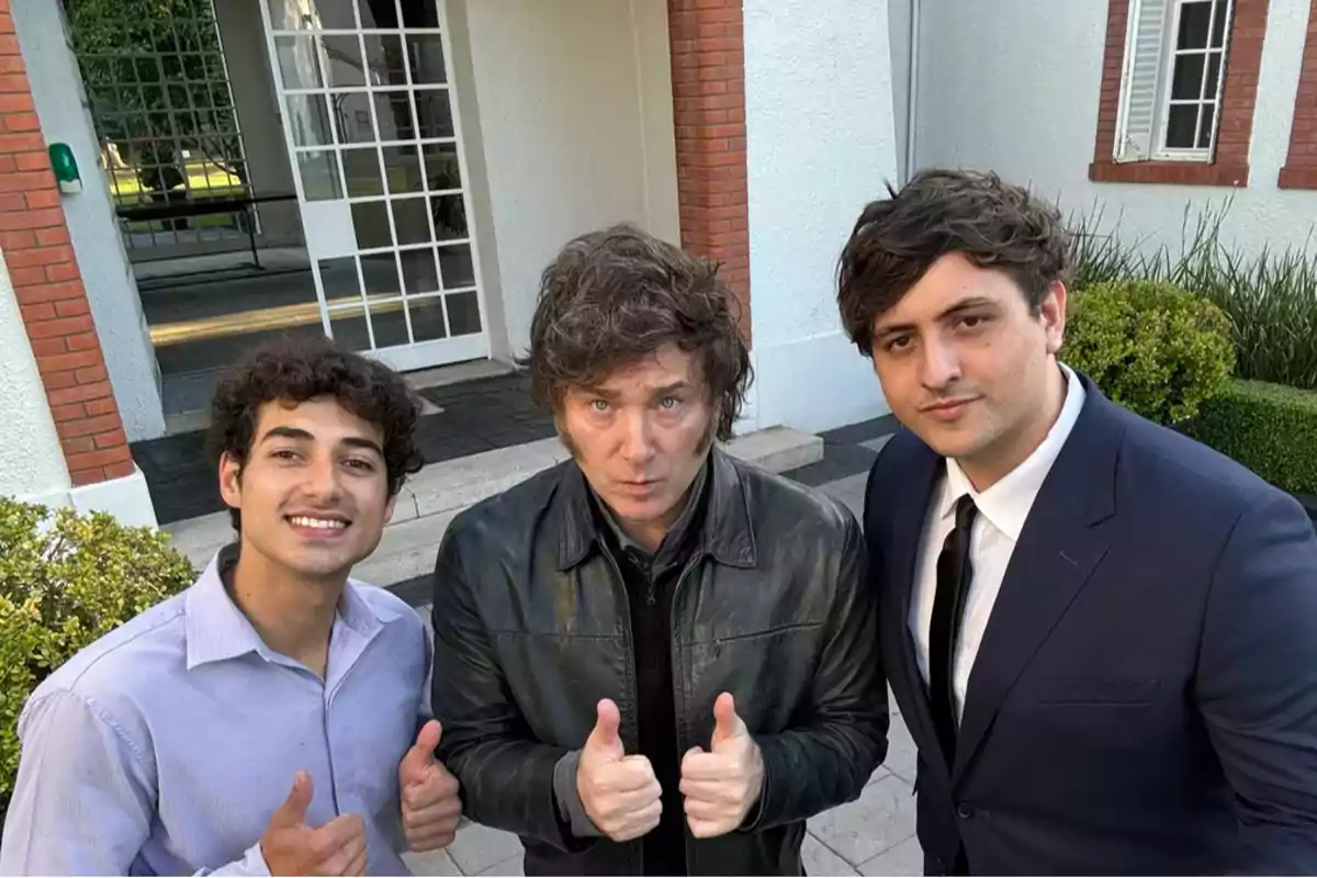 Tres personas posan juntas al aire libre frente a un edificio, dos de ellas levantan los pulgares.