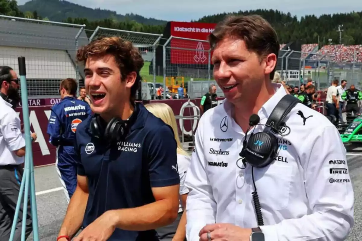 Dos personas con uniformes de Williams Racing sonríen en un circuito de carreras.