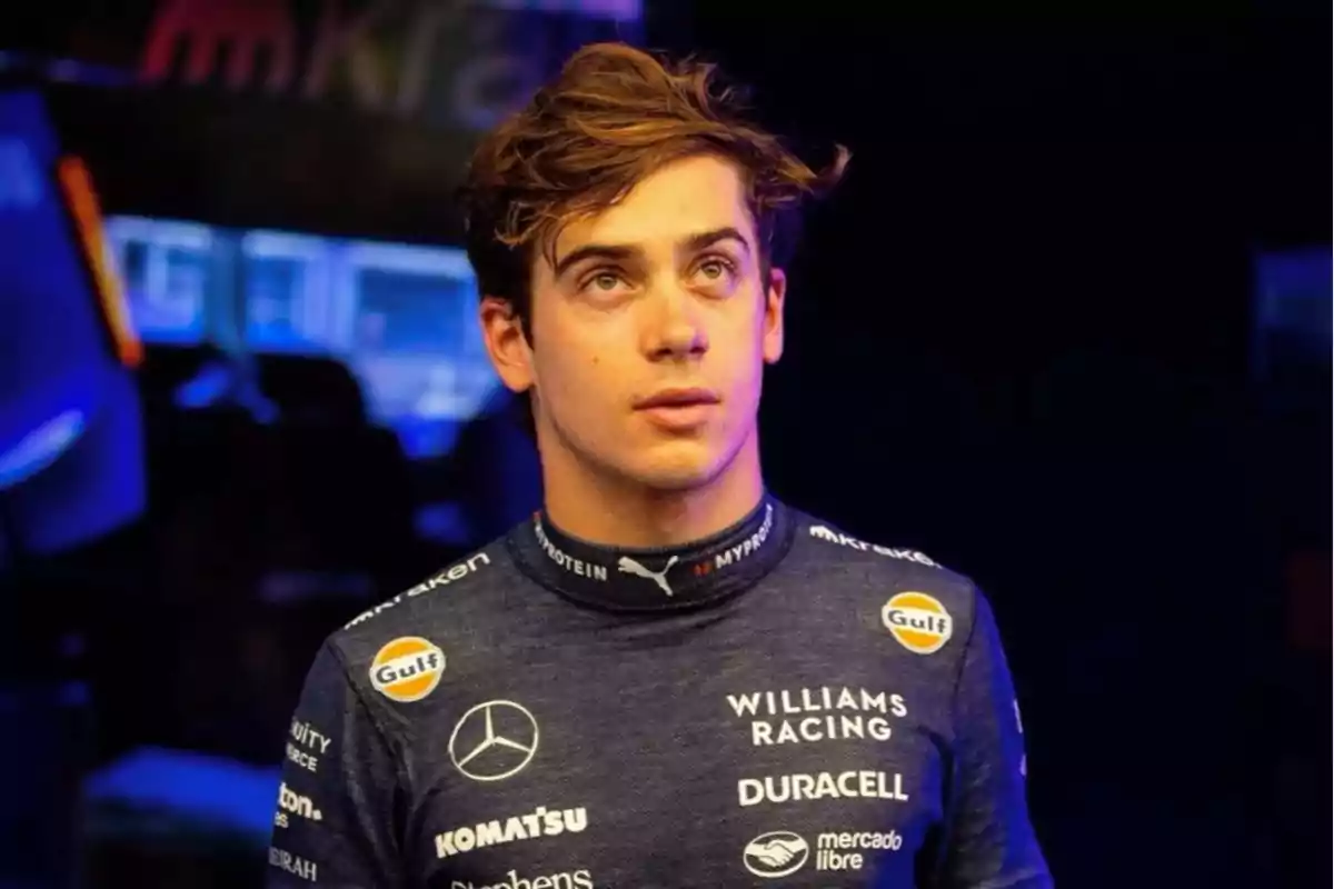 A race car driver in a dark blue Williams Racing uniform, with sponsor logos like Mercedes, Duracell, and Gulf, looking up in a lit environment.