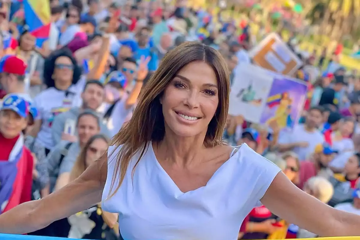 Una mujer sonriente en primer plano con una multitud de personas al fondo en un evento al aire libre.