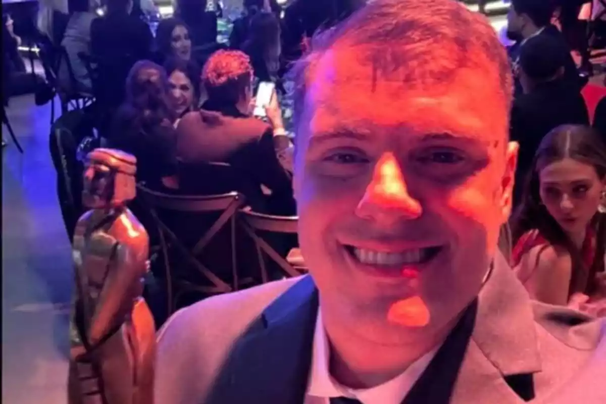 Un hombre sonriente en un evento formal sostiene un trofeo mientras otras personas están sentadas en el fondo.