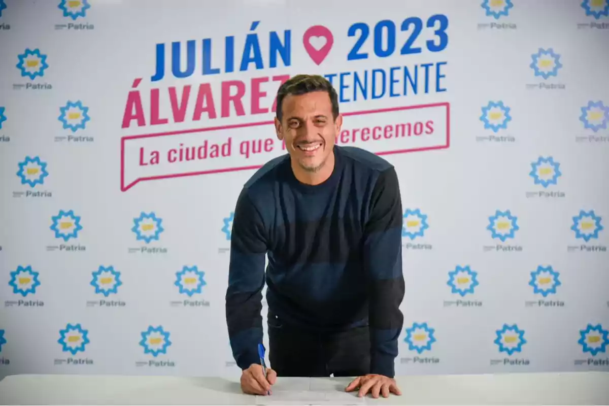 Un hombre sonriente firmando un documento frente a un cartel de campaña política que dice "Julián Álvarez 2023 Intendente, La ciudad que merecemos".