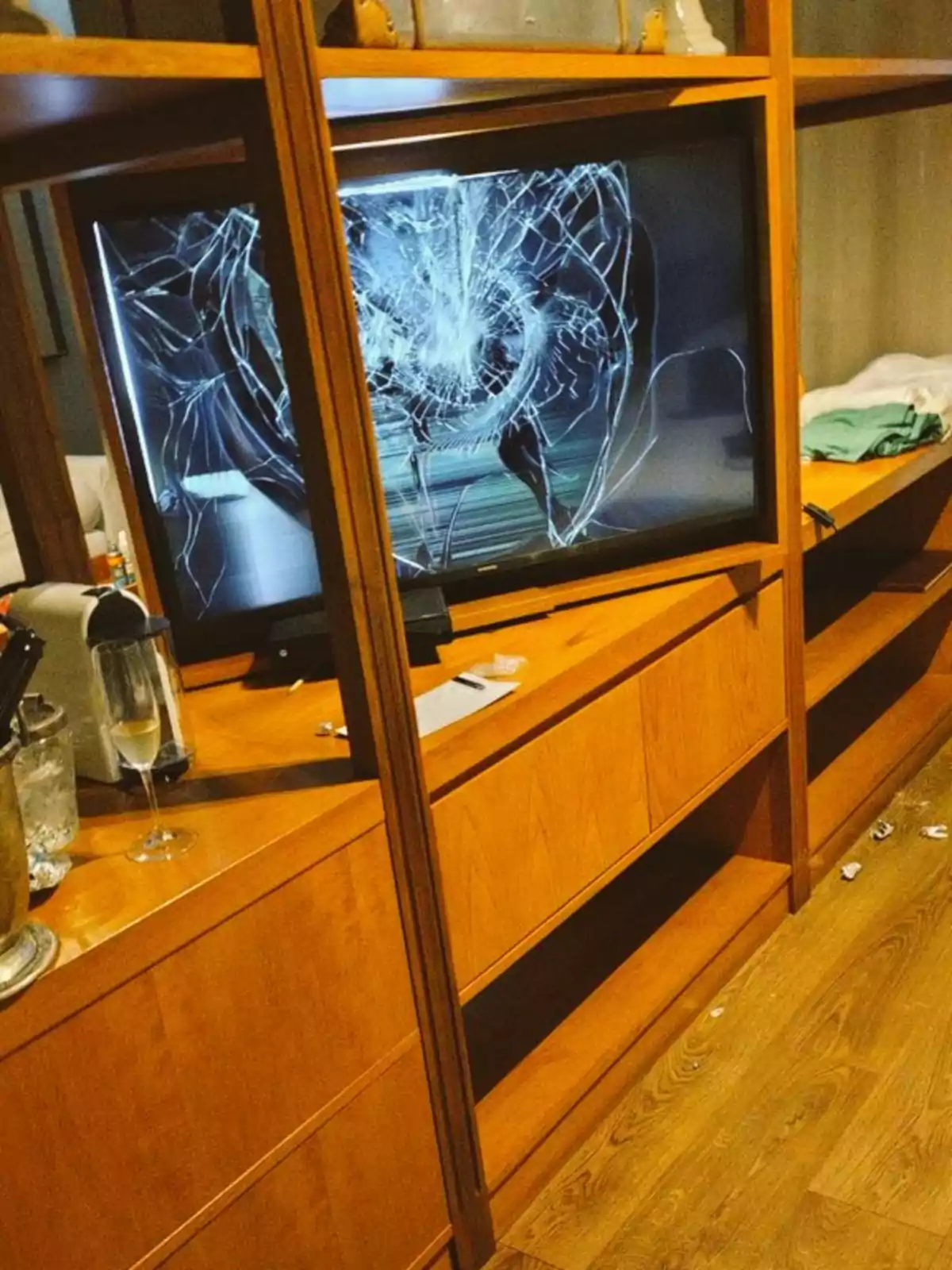 Un televisor con la pantalla rota se encuentra en un mueble de madera junto a una copa de champán y otros objetos.