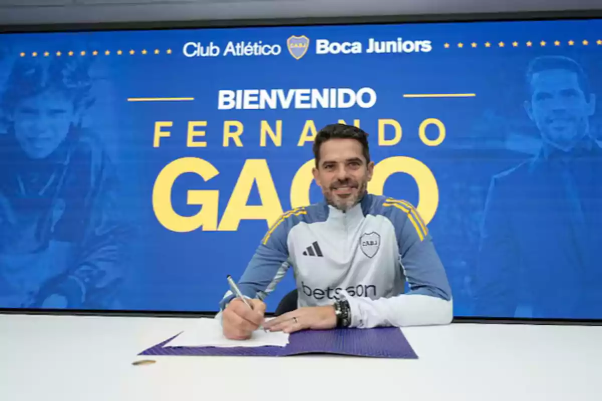 Un hombre firmando un documento frente a un cartel que dice "Bienvenido" y el nombre de un club de fútbol.