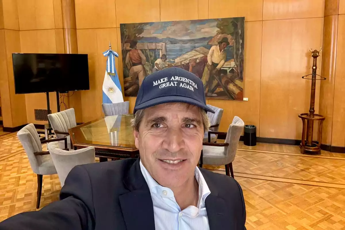 Un hombre con gorra azul que dice "Make Argentina Great Again" se toma una selfie en una sala de reuniones con una bandera argentina y una pintura al fondo.