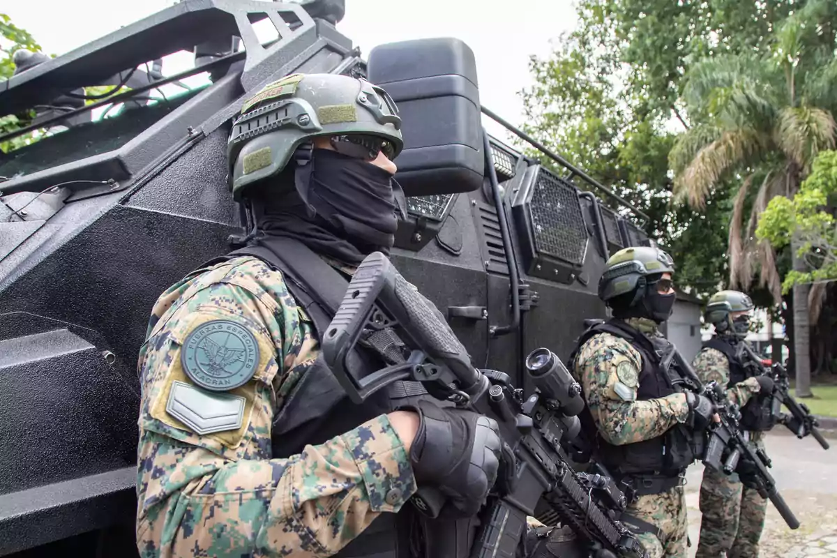 Soldados con equipo táctico y armas frente a un vehículo blindado en un entorno al aire libre.