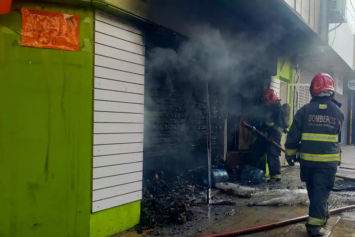 Bomberos trabajando para extinguir un incendio en un local comercial con humo saliendo de la entrada.