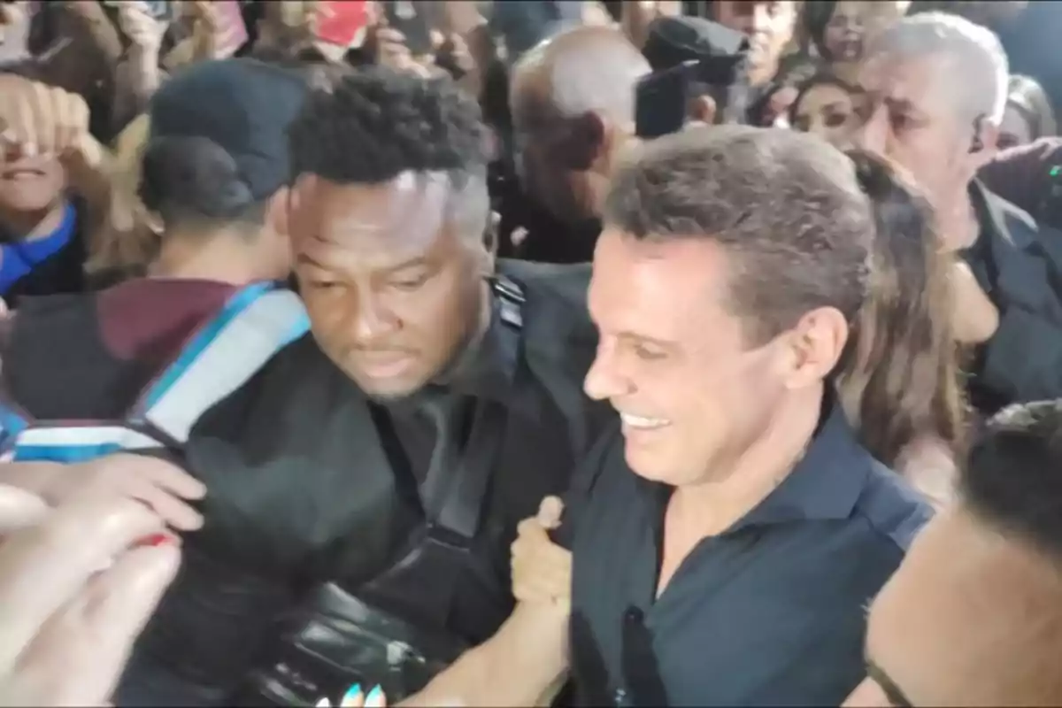 Un hombre sonriente rodeado de una multitud mientras es escoltado por un guardia de seguridad.