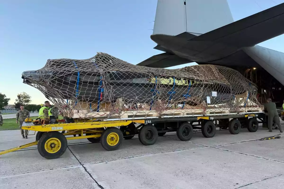 Un objeto grande cubierto con una red está siendo transportado en un remolque amarillo cerca de un avión militar en una pista de aterrizaje.