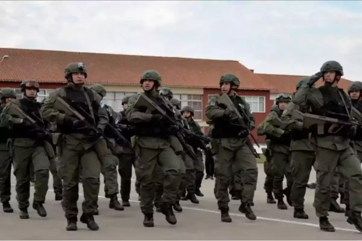 Soldados marchando en formación con uniformes y equipo militar frente a un edificio.