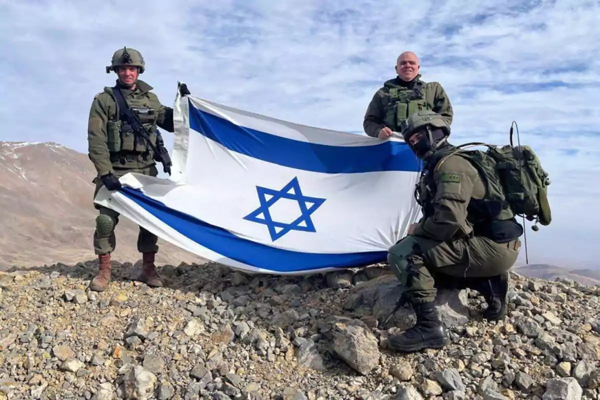 Tres personas con uniformes militares sostienen una bandera de Israel en un terreno rocoso con montañas al fondo.