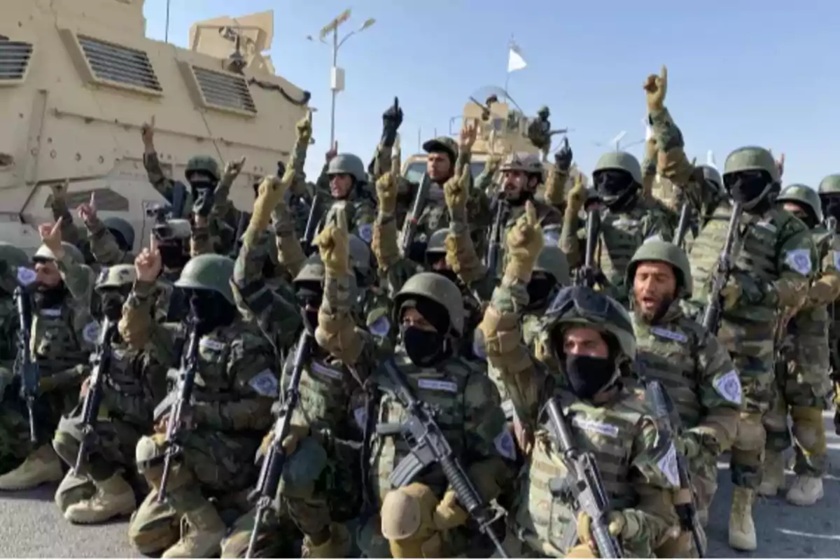 Un grupo de soldados en uniforme militar con cascos y armas levantando un dedo en señal de unidad frente a vehículos blindados.