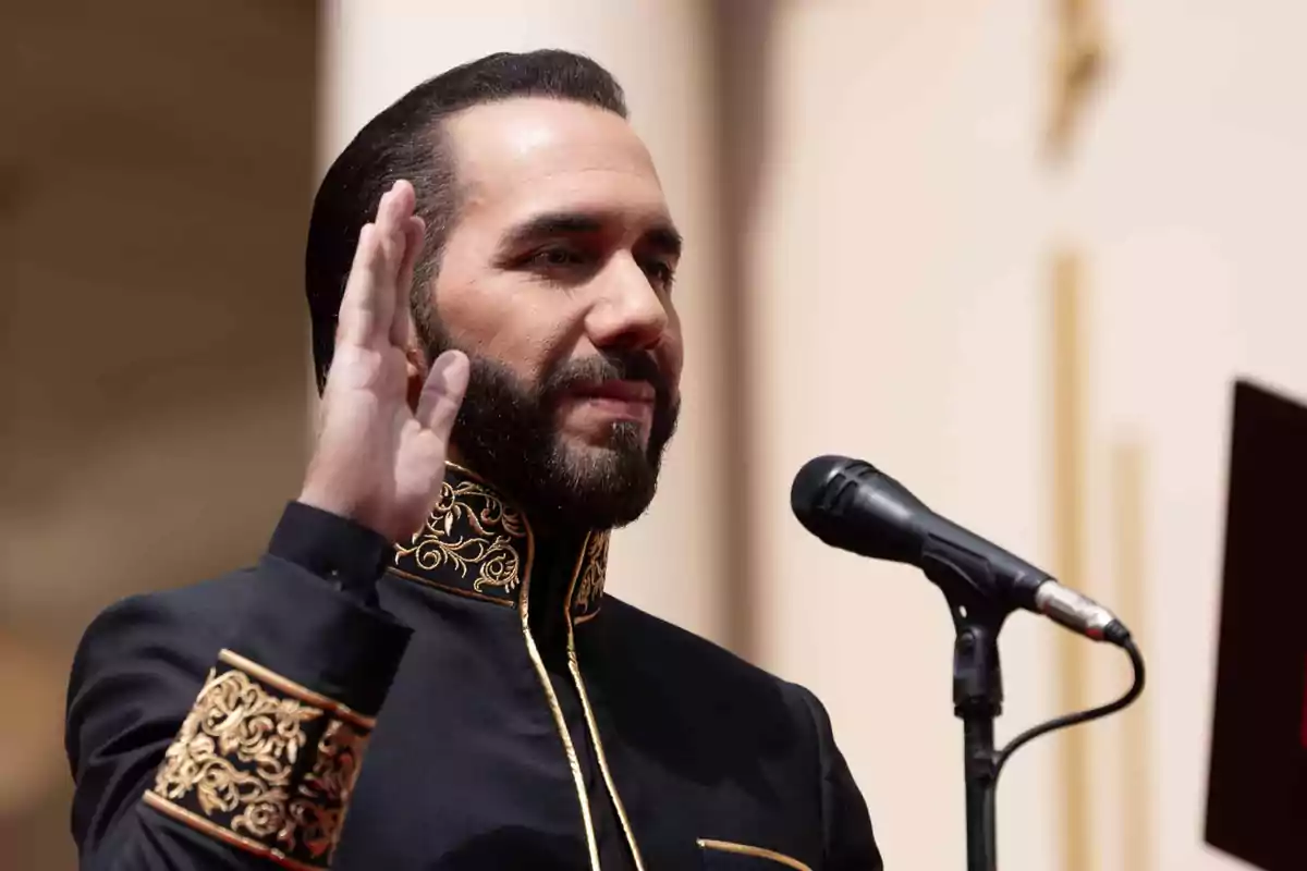 Un hombre con barba y cabello oscuro está frente a un micrófono levantando la mano derecha mientras lleva un traje negro con detalles dorados.