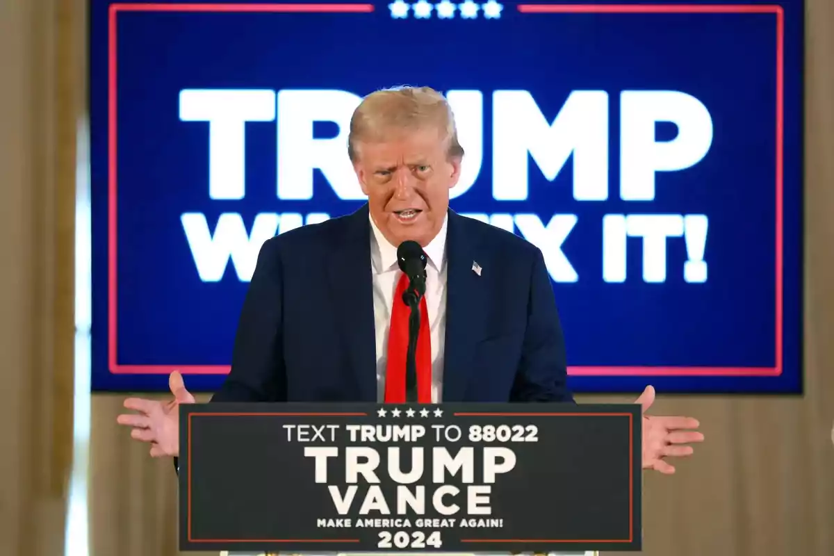 Un hombre de traje oscuro y corbata roja habla en un podio con un cartel que dice "Trump Vance 2024" y un fondo azul con texto blanco.