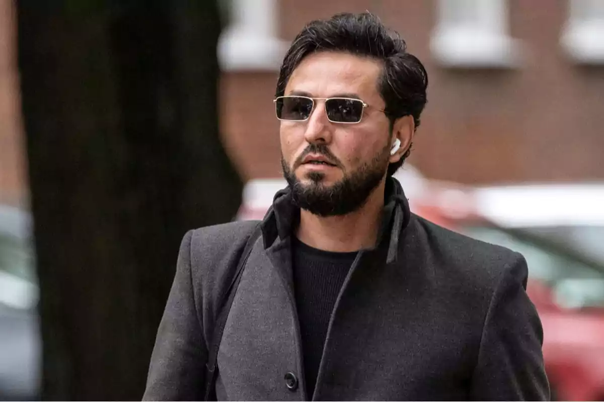 Un hombre con barba y gafas de sol camina por la calle usando auriculares y un abrigo oscuro.