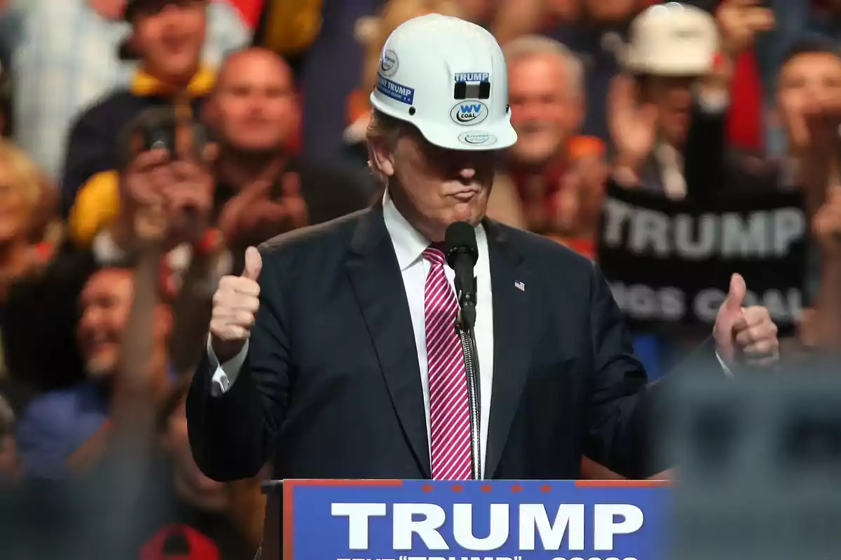 Un hombre con casco de construcción y traje oscuro, con corbata a rayas, habla en un mitin político mientras hace un gesto de aprobación con ambas manos; detrás de él, se ven personas sosteniendo carteles de apoyo.