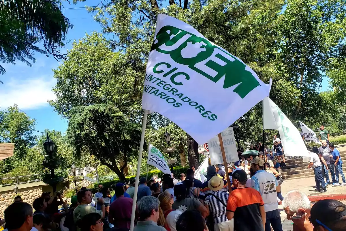 Un grupo de personas se reúne al aire libre sosteniendo banderas blancas con texto verde en un entorno arbolado.