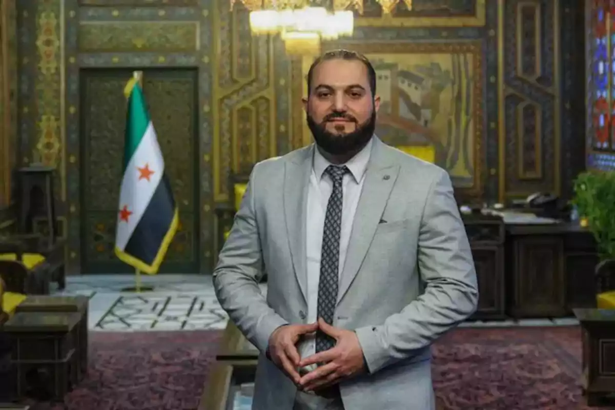 Un hombre con traje gris y corbata posa en una sala decorada con un estilo tradicional y una bandera con estrellas rojas al fondo.