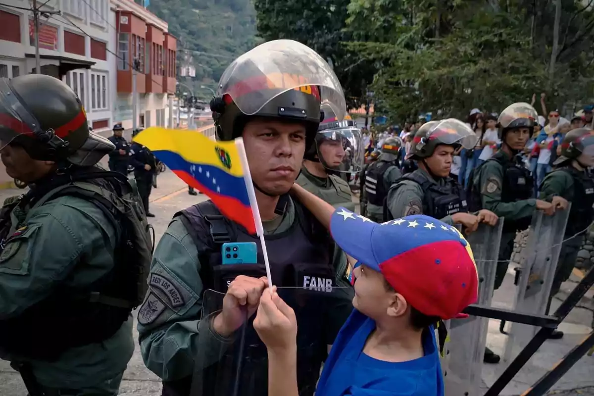 Un niño sostiene una bandera de Venezuela frente a un grupo de soldados con equipo antidisturbios.