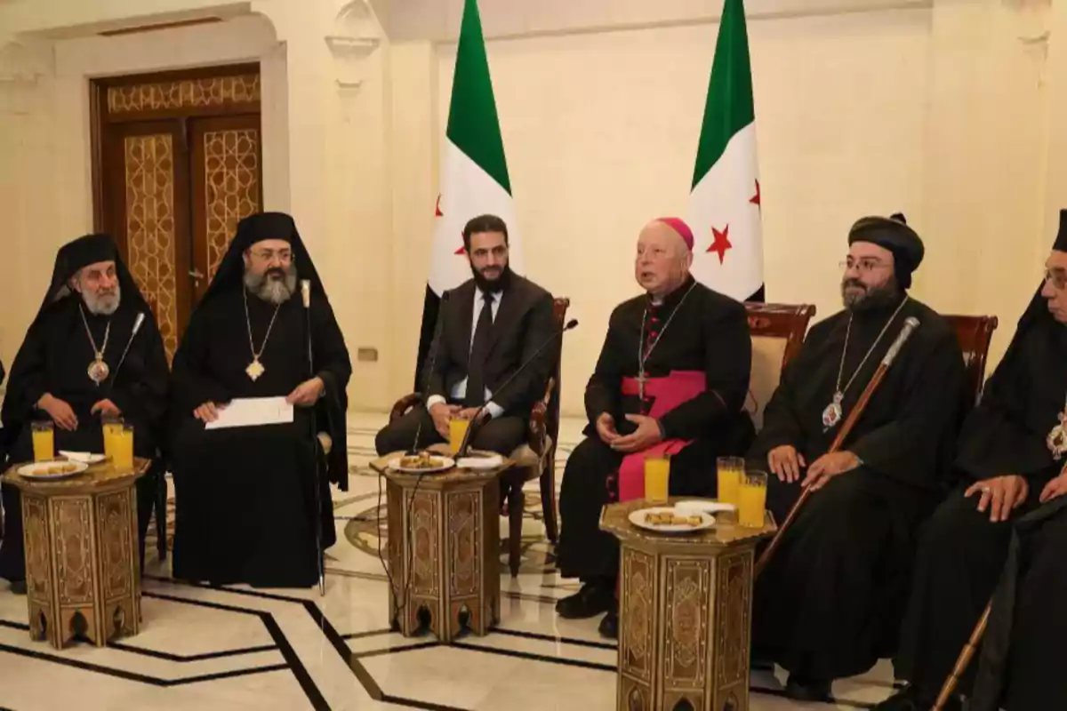 Un grupo de líderes religiosos y un hombre de traje están sentados en una sala con banderas de fondo y mesas con bebidas y platos frente a ellos.