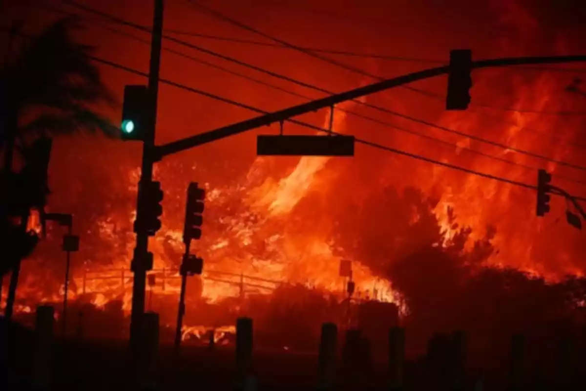 Incendio forestal ardiendo intensamente detrás de un semáforo en una intersección urbana durante la noche.