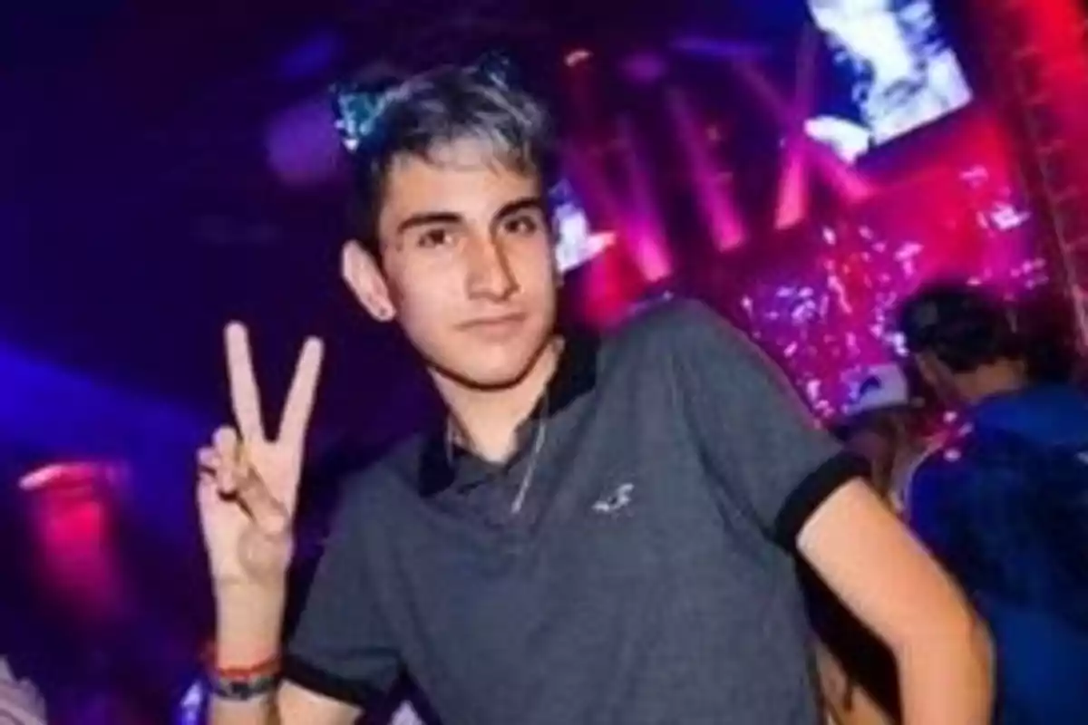 Un joven con orejas de gato y camiseta oscura hace el signo de paz en un club nocturno iluminado con luces de colores.