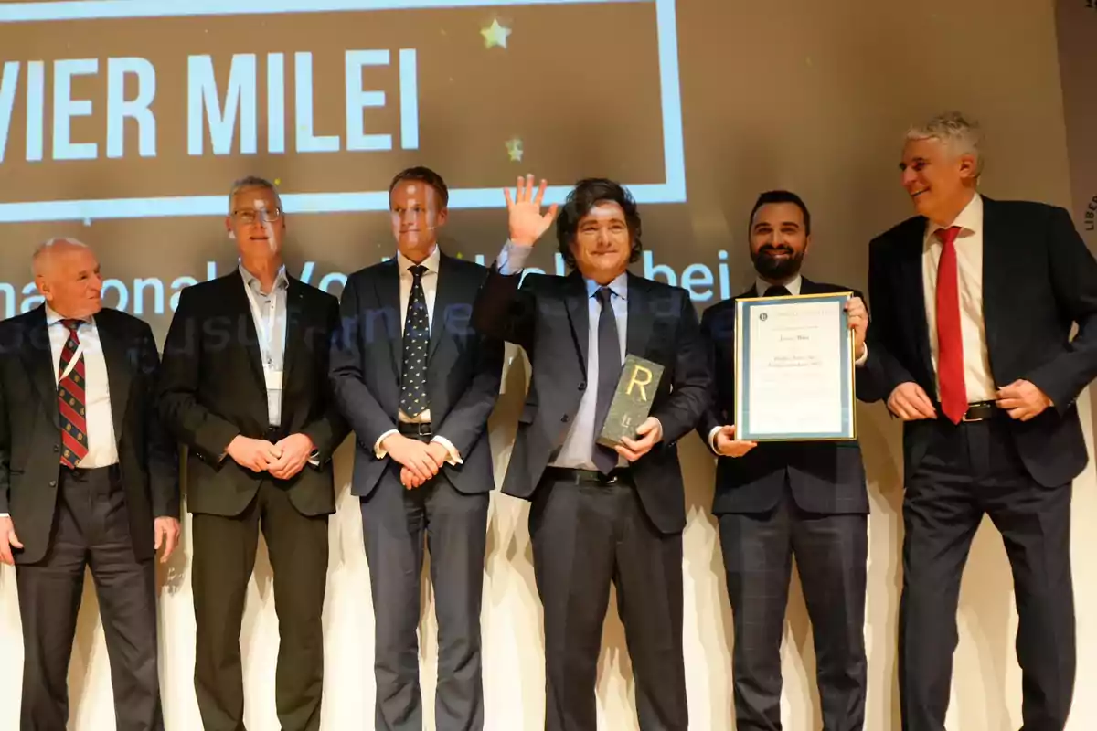 Un grupo de seis hombres en traje posando en un escenario, uno de ellos sostiene un trofeo y otro un certificado, con un fondo que muestra un nombre en letras grandes.